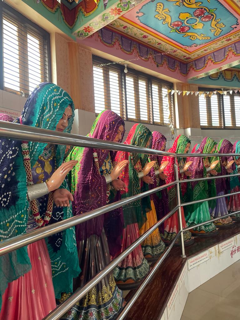 in the image some of the statues of women is present in multicoloured bandhani style odhni covering their head and wearing ghaghras. their right hand is raised to their chest as if to give blessings and their left nad is just under it.