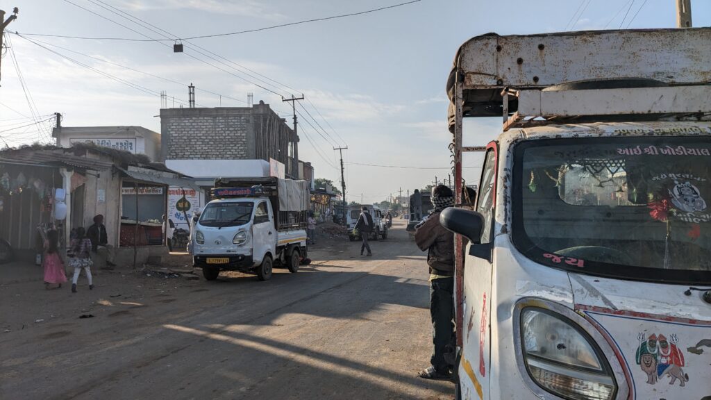 A scene at Balasar, Rapar. In the right side of the frame there is left front of a small pick-up truck. On the other side of the road, some people and small buildings and shops are present. A small pick up truck is also there.