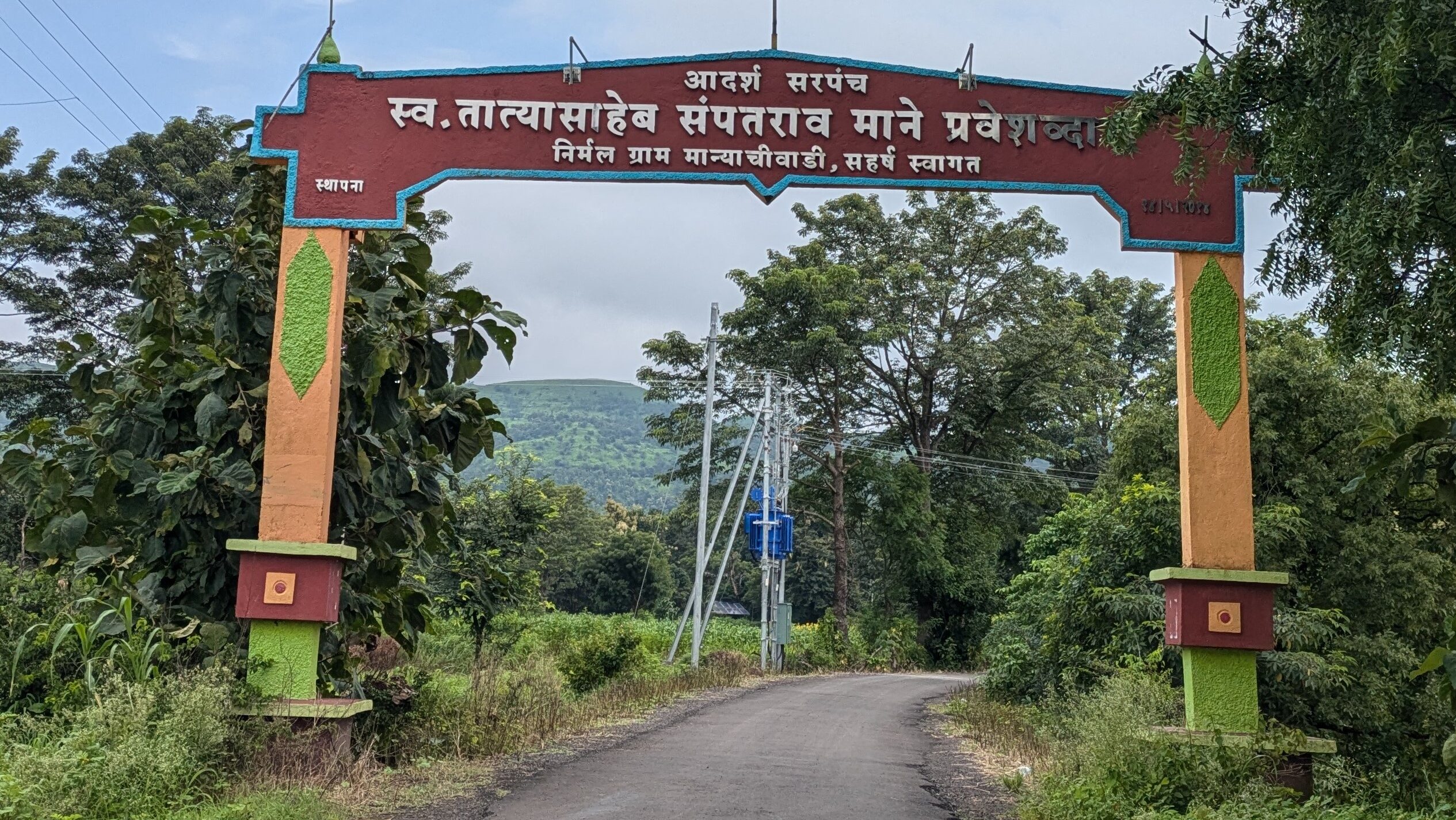 Entrance of the village. There's greenery on both sides of the road. An arch welcoming people stands on two pillars on either side of the road.