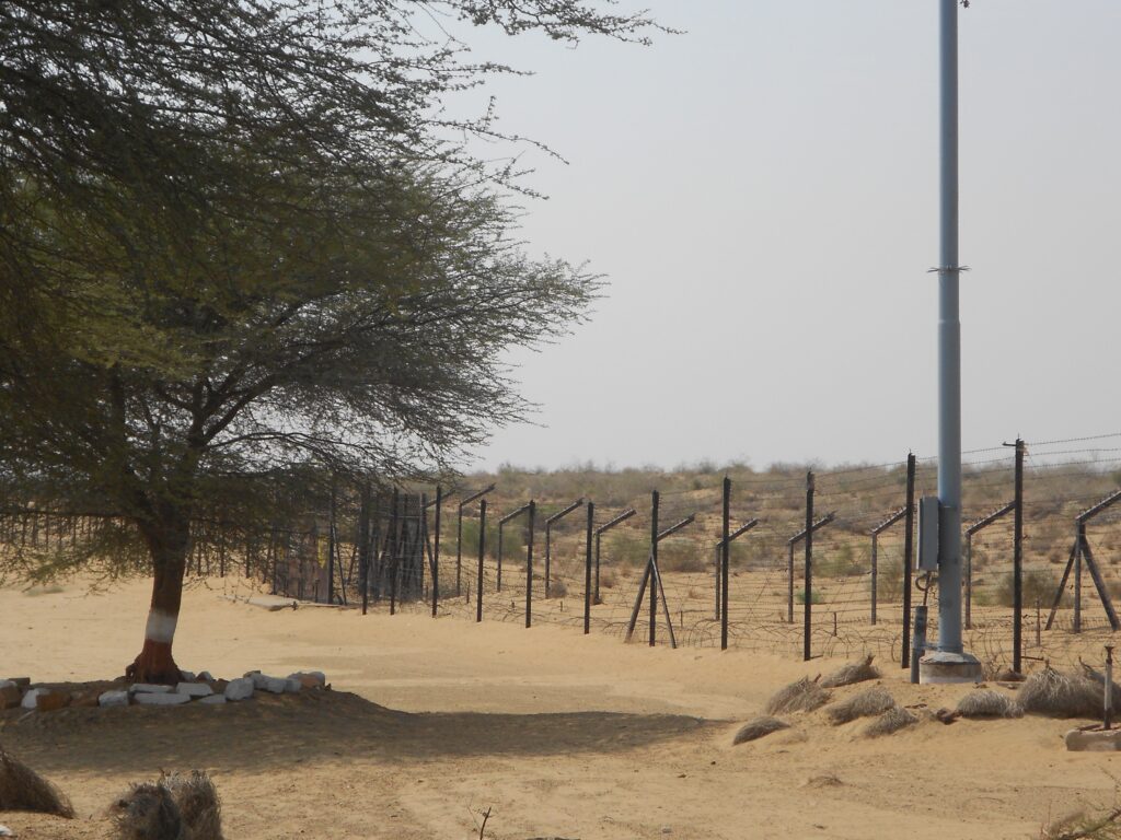 Image of a desolate land. There's a babool tree on the left, and barbed wire boundary on the right of the screen going towards left. Sky is visible in the two third of the right side.