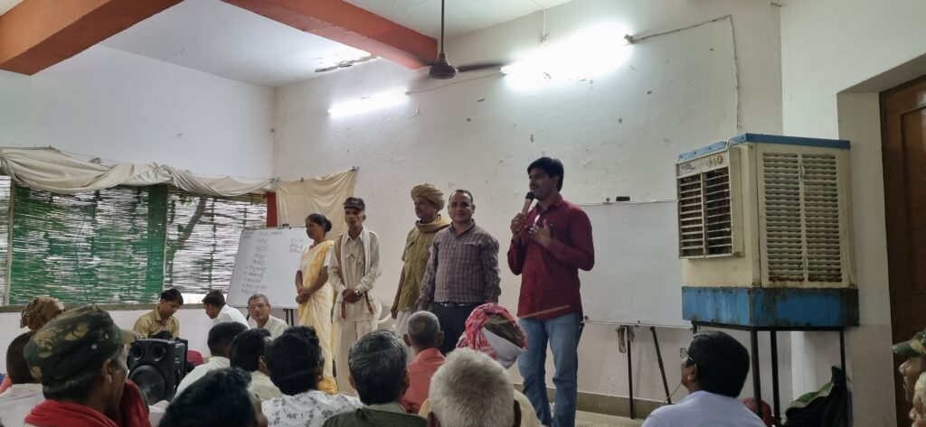 Four men wearing different clothes and a woman wearing saree are standing in a room with white walls, cooler to the right, and people sitting in front of them. The woman is to the extreme left, and the person on the extreme right is holding a mic and speaking. The picture has been taken so that the heads of the audience is in the frame from behind while the speakers fully appear in the centre. There are whiteboards to their either side with something written on the left one. To the left are three large windows with green chic covering them. The light in the room is largely from two overhead tubelights behind the speakers.