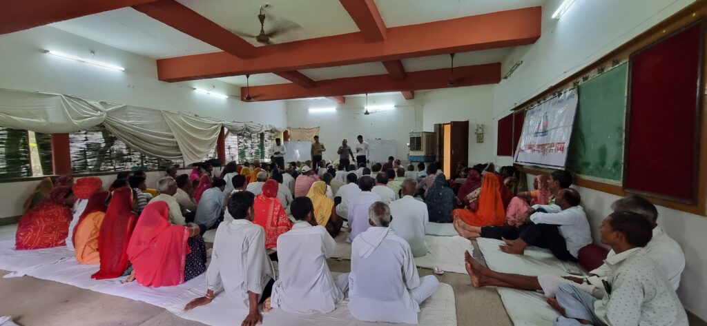 The image has been clicked from behind the audience. Many of them are sitting on mattresses covered with white sheet on ground and looking ahead. There are men and women of different ages in the audience. The room is large, with white walls and brown coloured beams on the roof. On the opposite end, three four persons are standing facing the audience. On the right side of the room there is a huge red rectangular pinboard with something pinned on it. To the left are large windows covered with green chic and white curtains, which are randomly up or down. The result of the union elections are being announced.