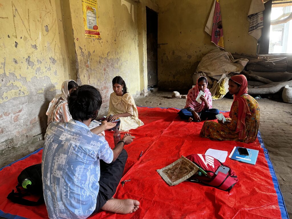 Aditya sits turned away from the camera and facing four women on a deep red carpet in a room with yellowed walls. There's some wood near the window.