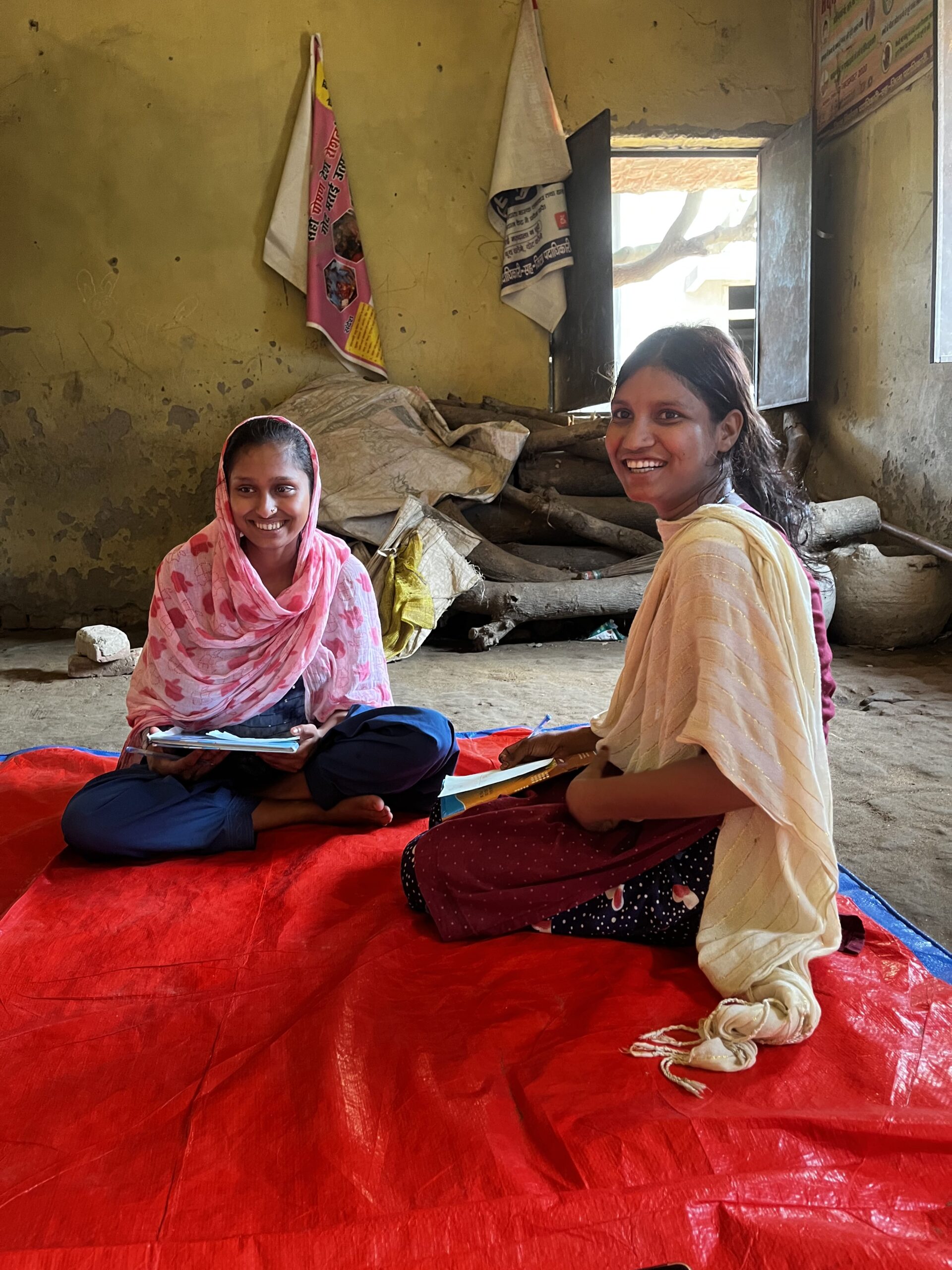 Two young women sitting on a deep red carpet look at the camera and smile. They are sitting in a room with yellowed walls and there is some wood near the window behind the women.