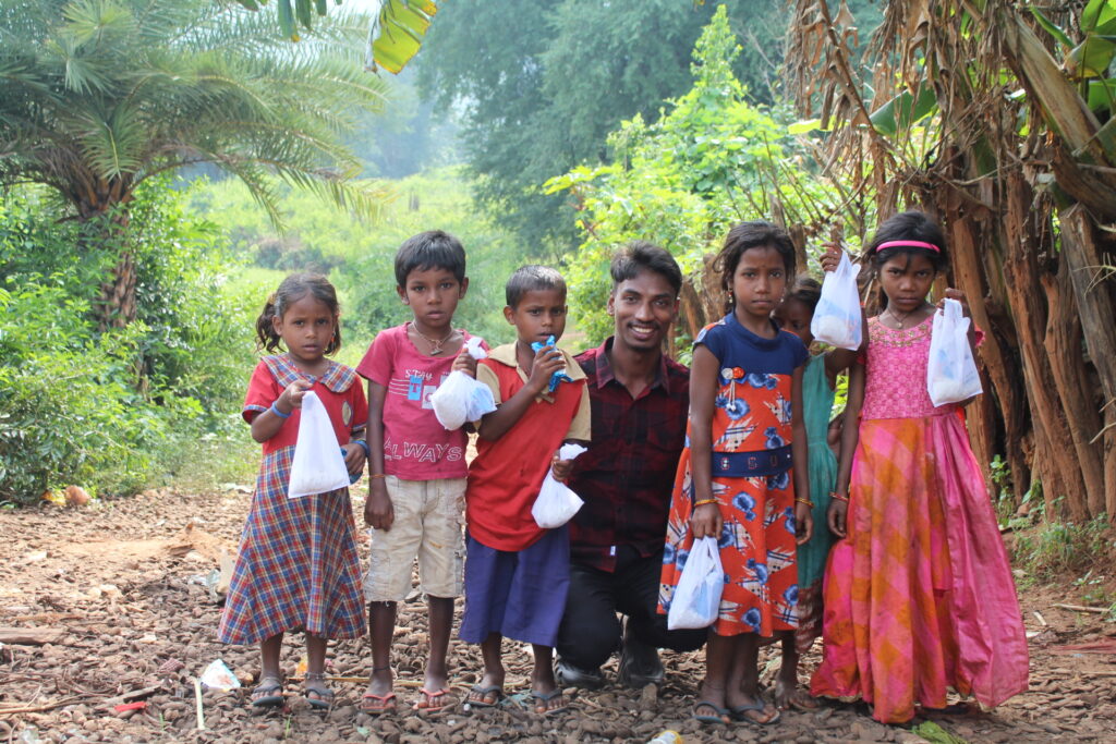 Rahul sits and poses with three children to his right and two to his left against trees, plants and shrubs green. The children are carrying something in a white polythene. Working with tribal healthcare involves great amount of work with children.