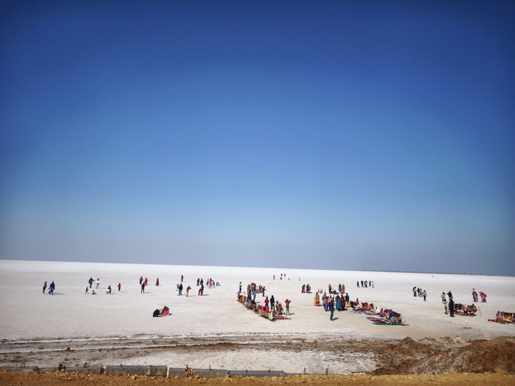 A landscape of white sand with people and shops covering below one third of the image. In the upper two third is blue sky.