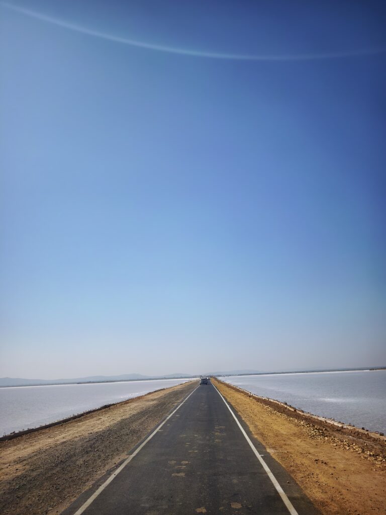 A portrait image of a road with white sand on both sides. The road appears to be meeting towards the far end. More than two third of the image is of blue sky.