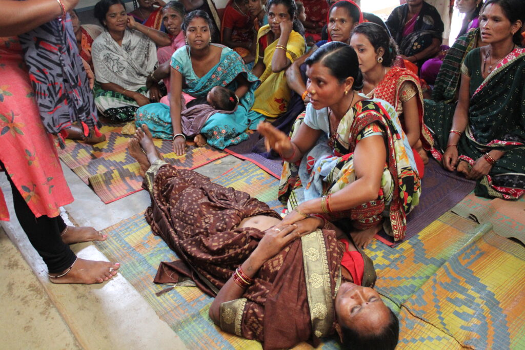 A woman in brown sari is lying down, another is sitting beside her and checking her pulse. Many other women are sitting behind them.