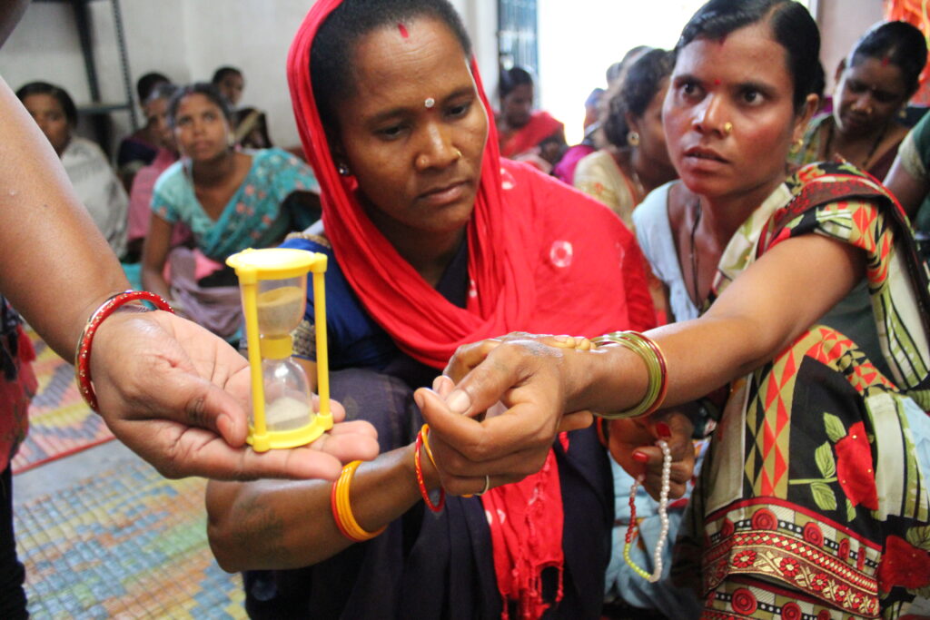 Image of a woman in red dupatta over her head sitting and checking the pulse of another woman to her left. Another hand to her holding hourglass is partially visible in the image. Several other women are sitting behind them.