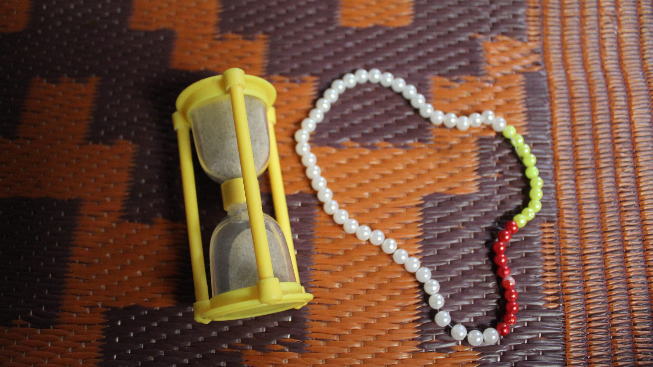 A small yellow hourglass with sand a beaded rosary is kept on a light brown plastic mat with dark brown geometrical shape. The rosary has largely white beads with a few yellow and red ones to measure breathing.