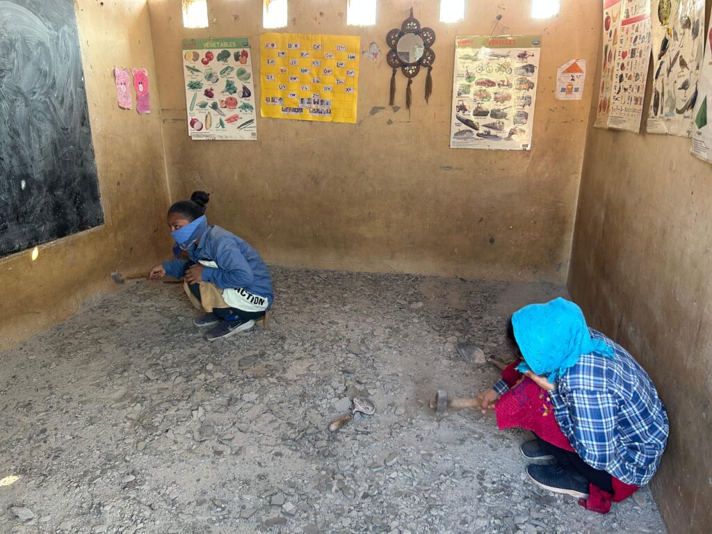 Picture of a small room with two girls sitting at either ends, one to the right of the image in front and other behind to the left end. The walls are light brown with some charts, one wall has a blackboard of which only a small section is visible. The floor is greyish, perhaps made of cement and sand.