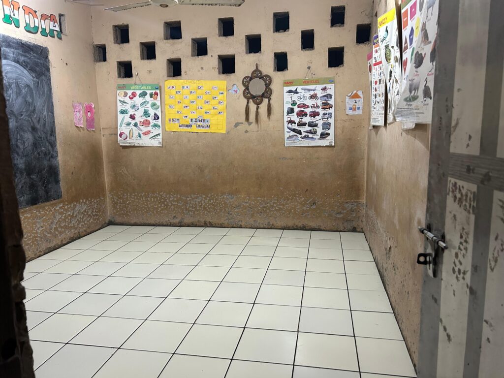 Picture of a small room. The walls are light brown with some charts and patterned square holes for ventilation, one wall has a blackboard of which only a small section is visible. The floor is tiled.