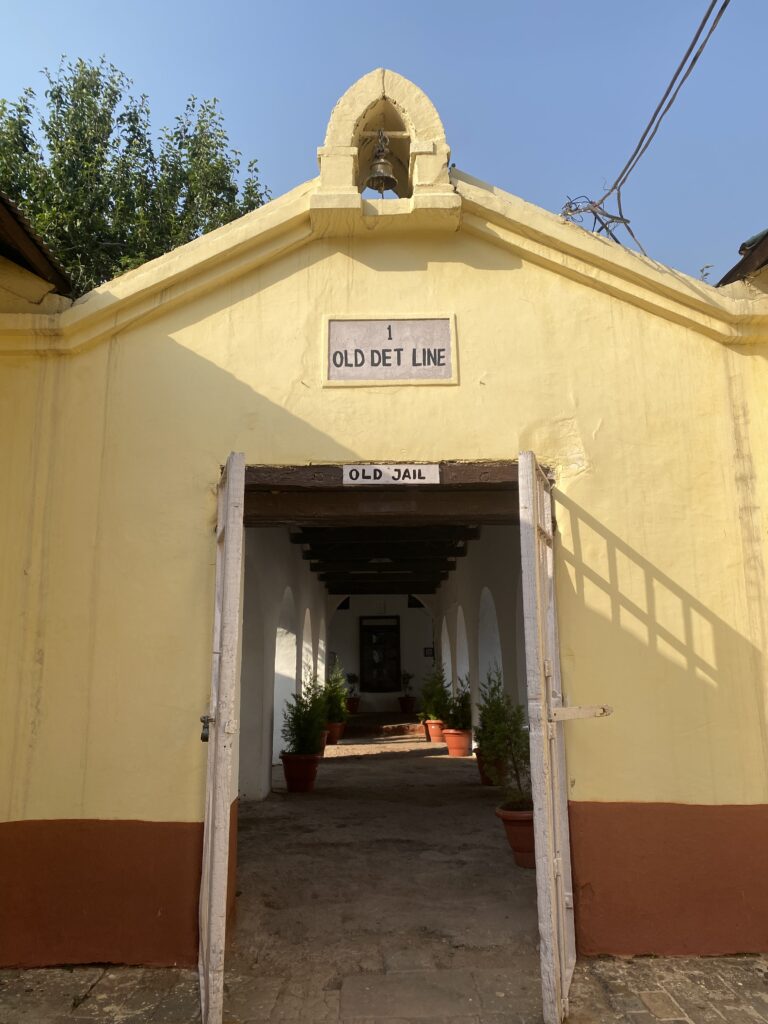 The entrance to Dagshahi jail. A cream yellow outer wall with brown paint from ground to upto 3 feet. The entrance seems open.