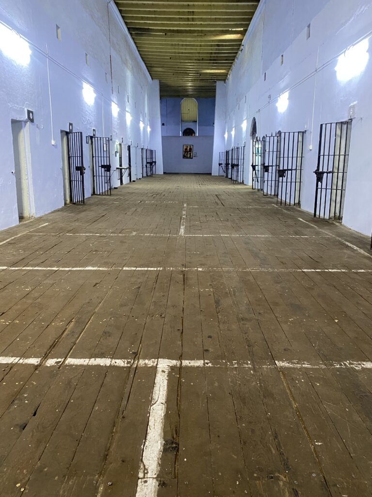A brown pathway leading upto a wall. On both sides of the pathway are walls with black gates opening outside. These are barracks. Wall is painted light blue.