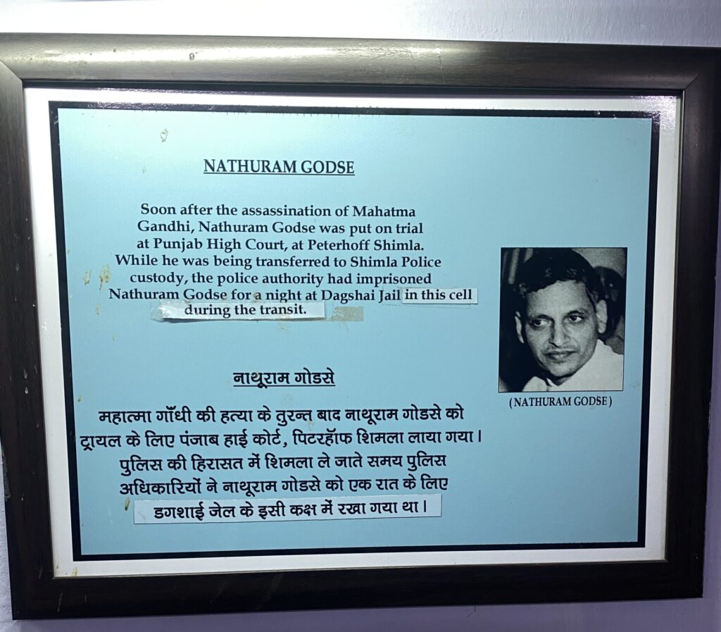 A plaque with Nathram Godse's picture to the right and text in Hindi and English stating that he was held here for transfer after he murdered Mahatma Gandhi.