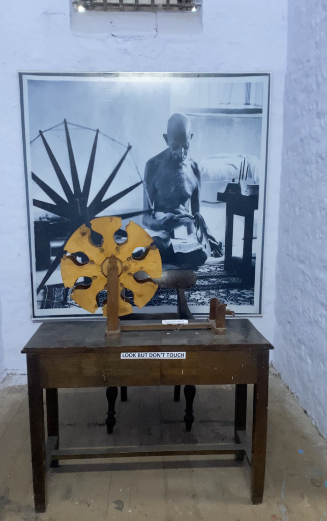 Gandhi's large portrait is hung on the blue wall- he is working on his charkha. A charkha is kept on a brown wooden table which reads: "Look but don't touch."